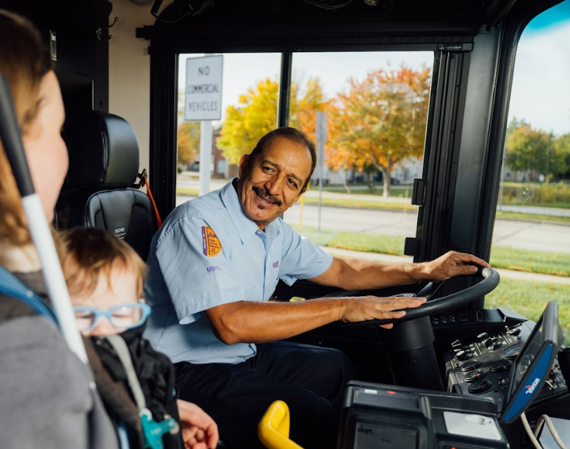 A smiling DART bus driver in uniform seated at the wheel, greeting a woman and young child boarding the bus