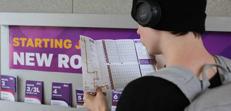 A person wearing headphones reading a DART bus route schedule brochure in front of a sign announcing new routes